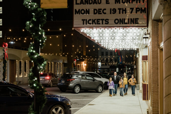 A family of five walks under the glowing marquee of the Granada Theater in downtown Bluefield at night. Christmas garland, twinkling string lights, and decorated lamp posts line the street, creating a festive holiday atmosphere. Cars are parked along the sidewalk as the family heads toward the theater entrance.