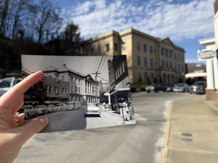 A historic black and white photo of a downtown street is held in front of the same location today, highlighting changes in buildings, cars, and streetscape over time.