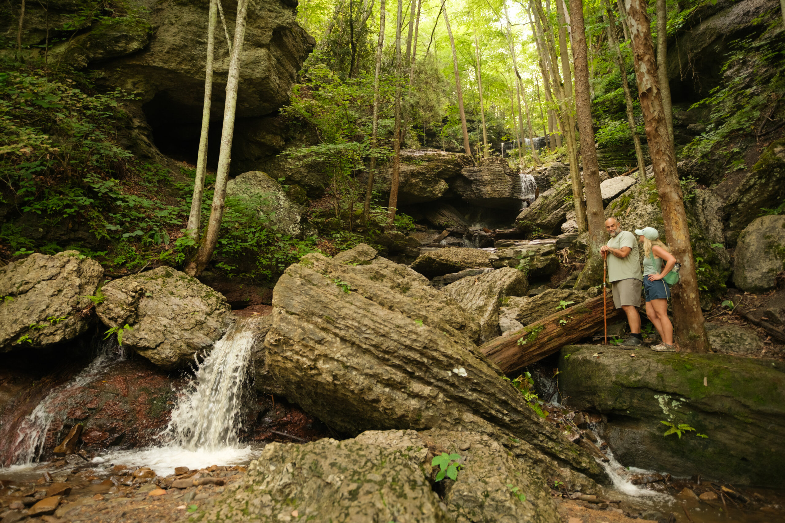 Two hikers stand on large rocks beside a small cascading waterfall in a forested rocky gorge surrounded by tall trees and moss-covered boulders.