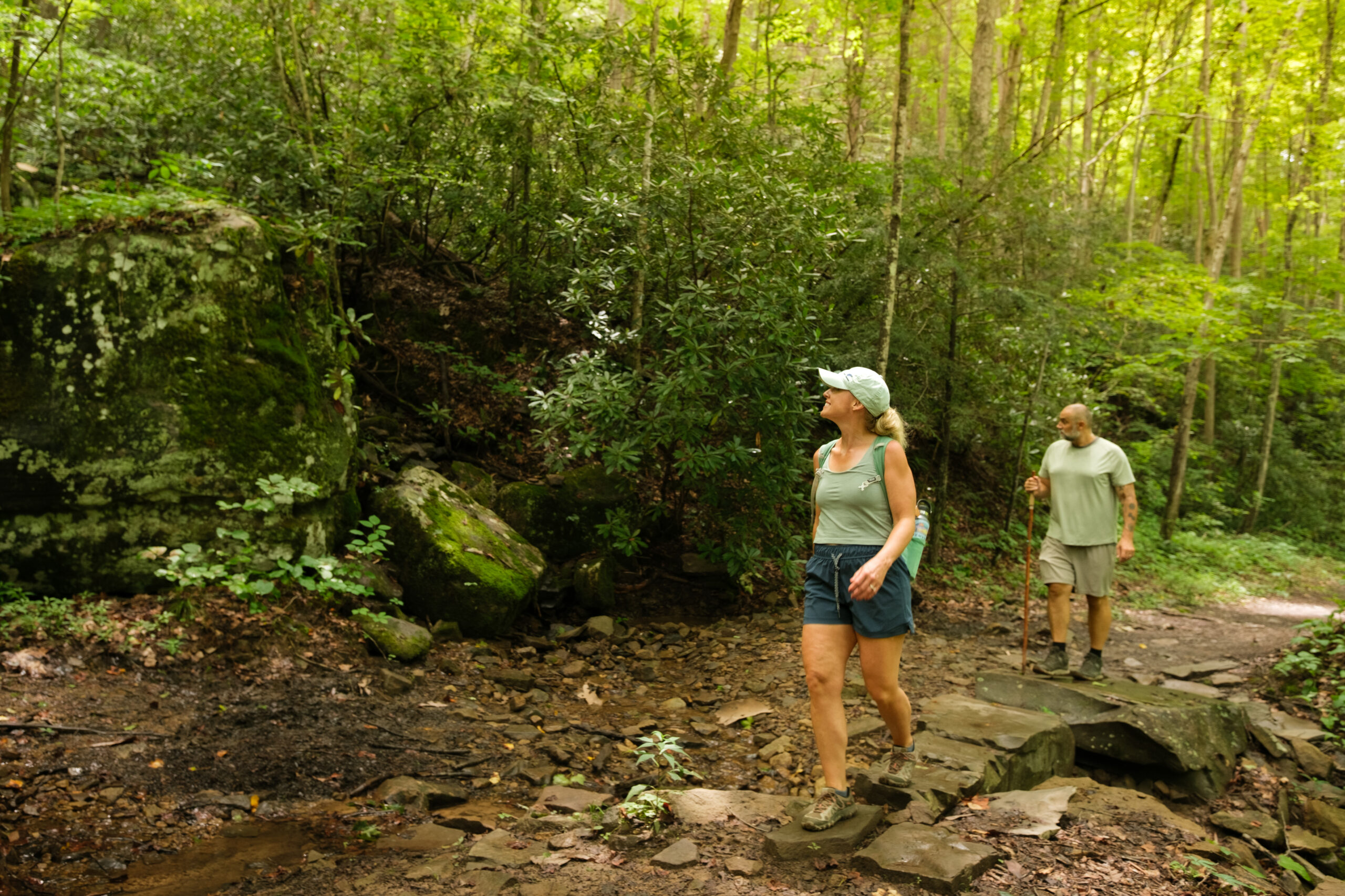 Two hikers walk along a forest trail, stepping across rocks beside a shallow stream surrounded by mossy boulders and dense green trees.