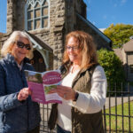 Two women stand outside a historic stone church in Bramwell, West Virginia, smiling as they look at a pink “Historic Bramwell” brochure. The church features a tall stone tower with arched stained-glass windows, and autumn trees and a stone building are visible in the background behind a black iron fence.