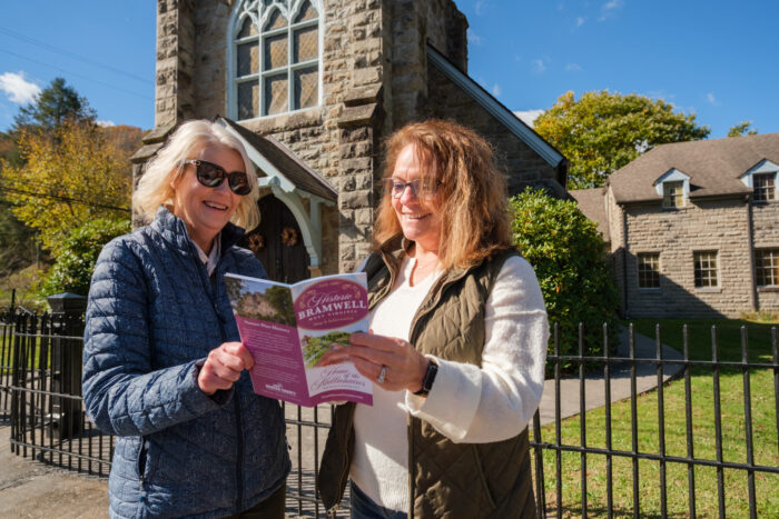 Two women stand outside a historic stone church in Bramwell, West Virginia, smiling as they look at a pink “Historic Bramwell” brochure. The church features a tall stone tower with arched stained-glass windows, and autumn trees and a stone building are visible in the background behind a black iron fence.