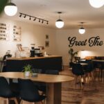 Interior of Aubade Coffee in downtown Princeton, West Virginia, featuring a modern black coffee bar with espresso equipment, wooden tables and black chairs, globe pendant lighting, a small seating area with a leather couch, and a wall mural that reads “Greet The Dawn.”