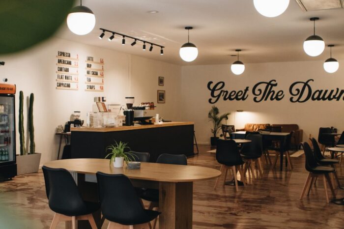 Interior of Aubade Coffee in downtown Princeton, West Virginia, featuring a modern black coffee bar with espresso equipment, wooden tables and black chairs, globe pendant lighting, a small seating area with a leather couch, and a wall mural that reads “Greet The Dawn.”