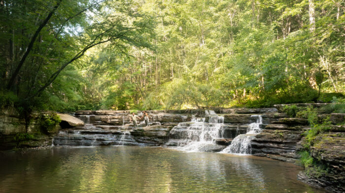 A wide view of a layered waterfall flowing into a calm pool surrounded by dense green forest. Two people stand on the rock ledges near the center of the falls, holding hands as sunlight filters through the trees above.