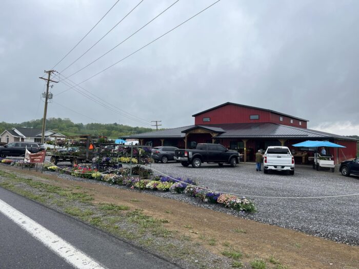 Exterior view of Market Over Yonder, a large red barn-style market with a black metal roof, seen from the roadside on an overcast day. Colorful hanging baskets, flowering plants, and garden displays line the front entrance while several vehicles are parked in the gravel lot. Rolling green hills and utility lines are visible in the background.