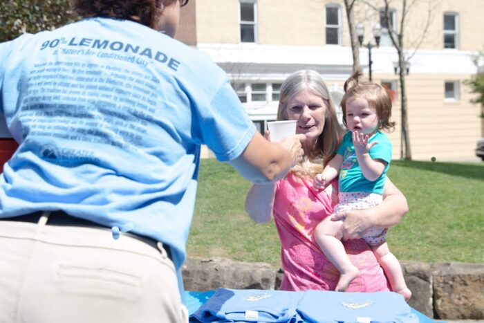 A volunteer wearing a light blue "90° Lemonade – Nature's Air Conditioned City" T-shirt hands a cup of free lemonade to a woman holding a young child at an outdoor table in Bluefield, West Virginia. Folded event T-shirts sit on the table, with historic downtown buildings and a sunny park in the background.