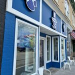 Exterior of Q38 on Mercer Street in downtown Princeton, West Virginia. The storefront features a bright blue facade with white trim, large front windows, baseball-themed logo signs, and white metal chairs along the sidewalk. An American flag hangs outside, with neighboring historic downtown buildings visible in the background.