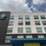 Exterior view of the Tru by Hilton hotel building featuring a modern white and black facade with blue and yellow accents at the main entrance.