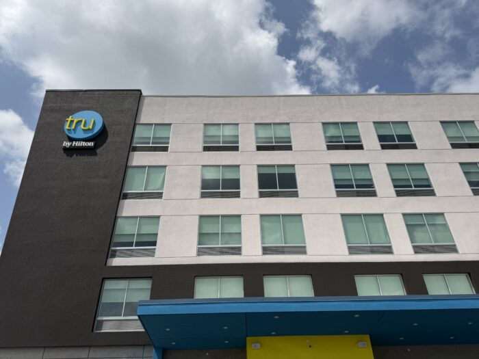 Exterior view of the Tru by Hilton hotel building featuring a modern white and black facade with blue and yellow accents at the main entrance.