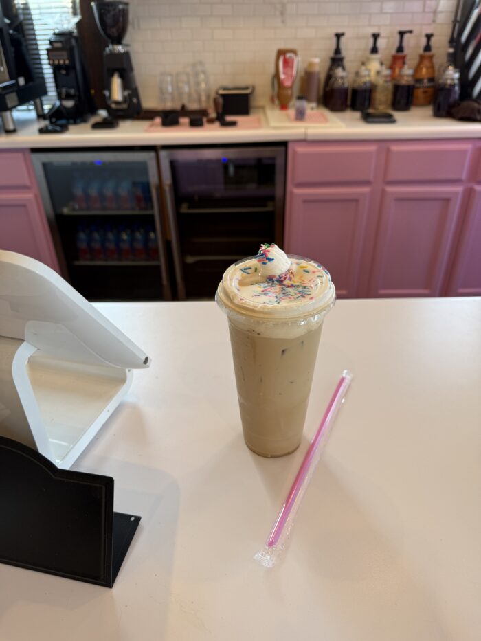 Iced coffee topped with whipped cream and colorful sprinkles sits on a white countertop inside a coffee shop with pink cabinets and espresso equipment in the background.